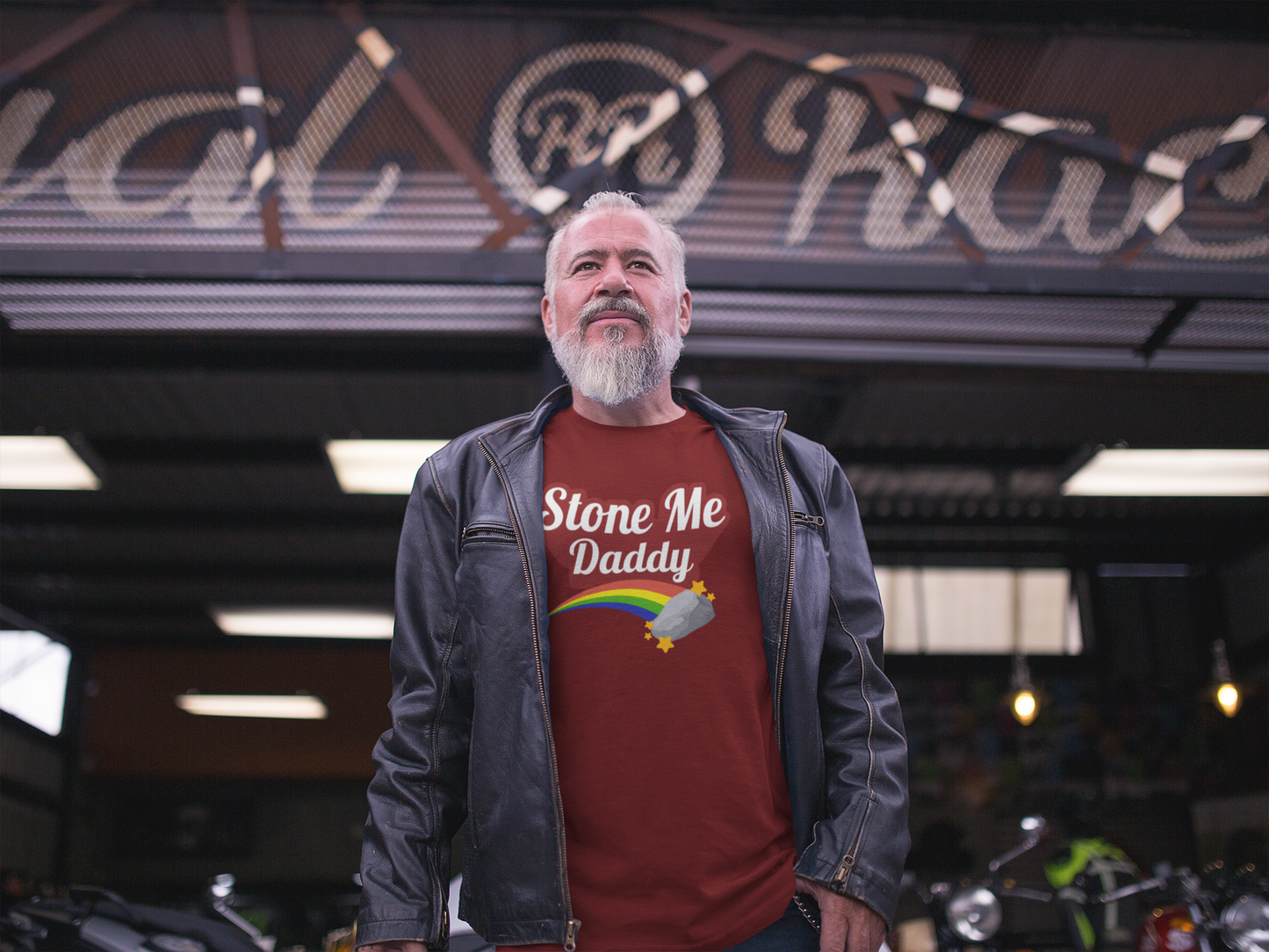 Person standing outside a motorcycle garage wearing a red “Stone Me Daddy” t-shirt with rainbow and stone graphic from Levitic*nts Apparel.